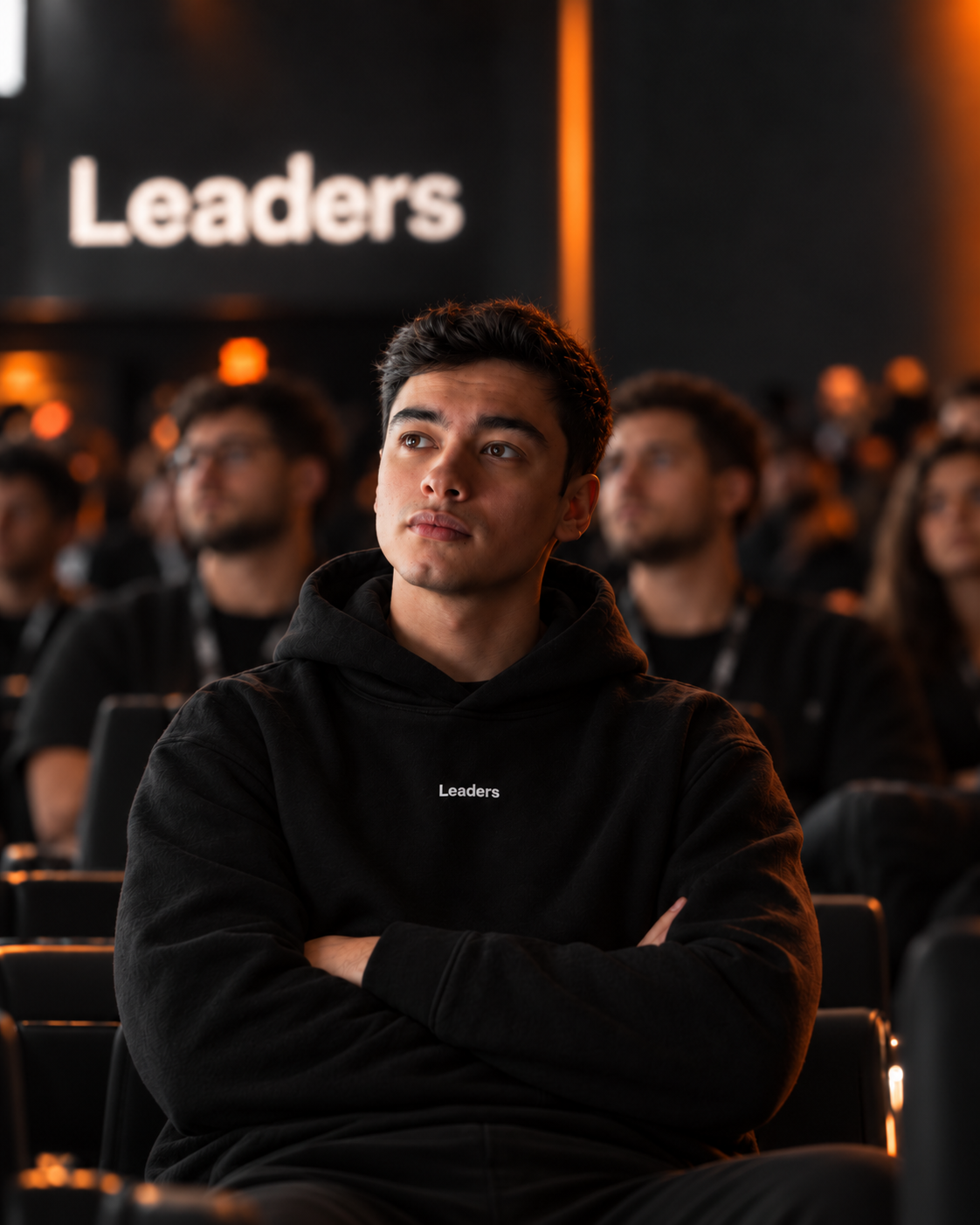 Bira sentado na plateia de um evento da Leaders, de moletom preto, com o letreiro da marca ao fundo.
